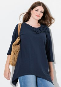 Mörkblå tunika med trekvartsärm, med texturerade blommor på bröstet. Kombinationen är med en vävd stråväska.