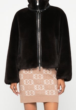 Femme portant une veste noire zippée en tissu duveteux avec col à boucle et une jupe beige à motif avec un design blanc entrelacé.