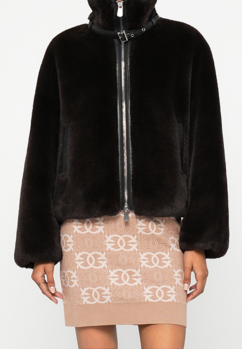 Femme portant une veste noire zippée en tissu duveteux avec col à boucle et une jupe beige à motif avec un design blanc entrelacé.