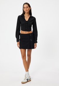 Cropped shirt nera con bottoni frontali e maniche lunghe, abbinata a una mini gonna nera con anelli laterali. La modella indossa scarpe da ginnastica bianche.