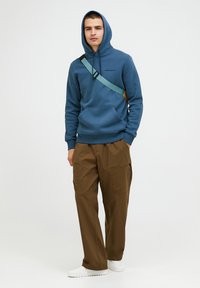 Felpa blu con cappuccio regolabile e tasca frontale, abbinata a pantaloni cargo marroni con tasche laterali e vestibilità comoda. Sneakers bianche.