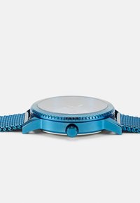 Reloj de pulsera metálico azul con esfera redonda, correa de malla texturizada azul y corona. Presenta una tapa de vidrio transparente y un diseño de esfera simple.