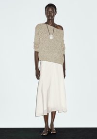 Beige gebreide trui met een losse pasvorm en een off-shoulder ontwerp, gecombineerd met een witte vloeiende maxi rok en eenvoudige sandalen.