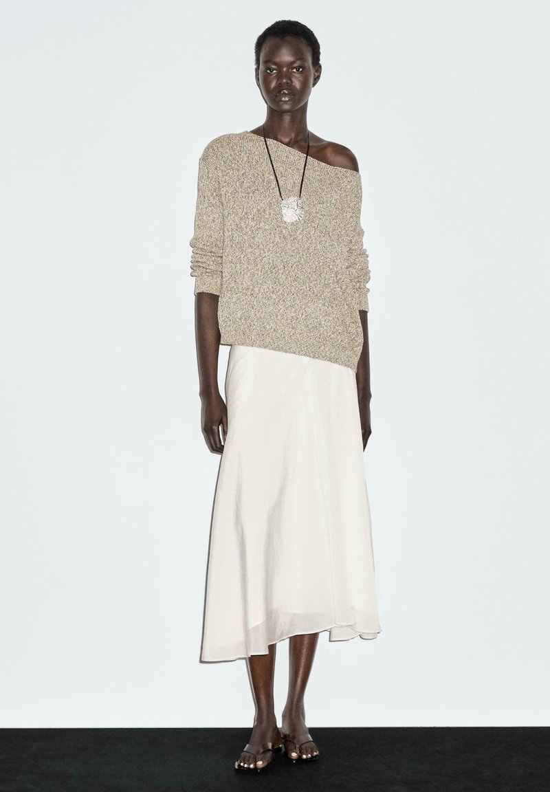 Beige gebreide trui met een losse pasvorm en een off-shoulder ontwerp, gecombineerd met een witte vloeiende maxi rok en eenvoudige sandalen.