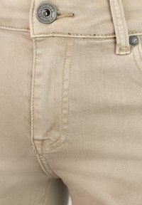Jeans de mezclilla beige con cierre de botón metálico, bolsillos cosidos y una tela suave y texturizada con un brillo sutil.