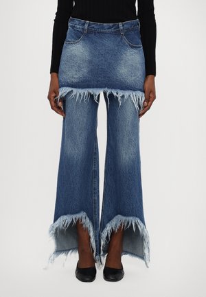 Modelka nosi niebieskie dżinsy z dołączoną mini spódniczką i postrzępionymi brzegami, zestawione z czarnymi szpiczastymi butami i czarnym topem z długim rękawem.