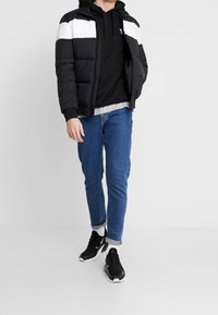 Giacca nera imbottita con strisce bianche sulle spalle, felpa nera con cappuccio, maglietta grigia, jeans blu e sneakers nere con accenti bianchi.