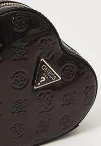 Svart hjerteformet veske laget av blankt materiale, med pregete mønstre og et triangelrødt gullsymbol med teksten "GUESS U.S.A.".