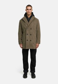 Olīvu zaļa divrindu mētelis ar melniem pogām, ar šalli, uzvilkts pāri melnai kapucē un melnām biksēm, stāvot priekšā vienkāršai fona.