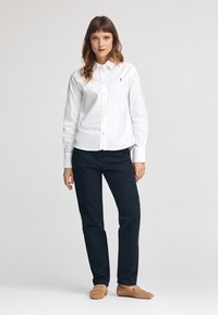 Camicia bianca a maniche lunghe con bottoni e un piccolo logo, abbinata a pantaloni scuri e scarpe beige senza lacci su uno sfondo neutro.