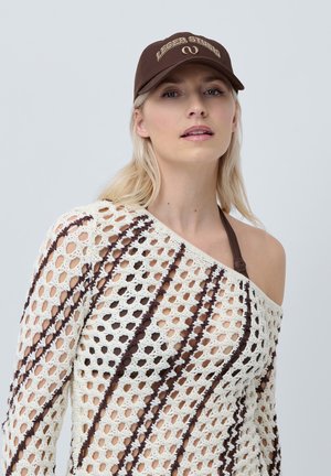 White knit sweater with brown stripes, featuring an asymmetrical design. A brown cap with "LEGER STUDIO" in gold lettering is worn.