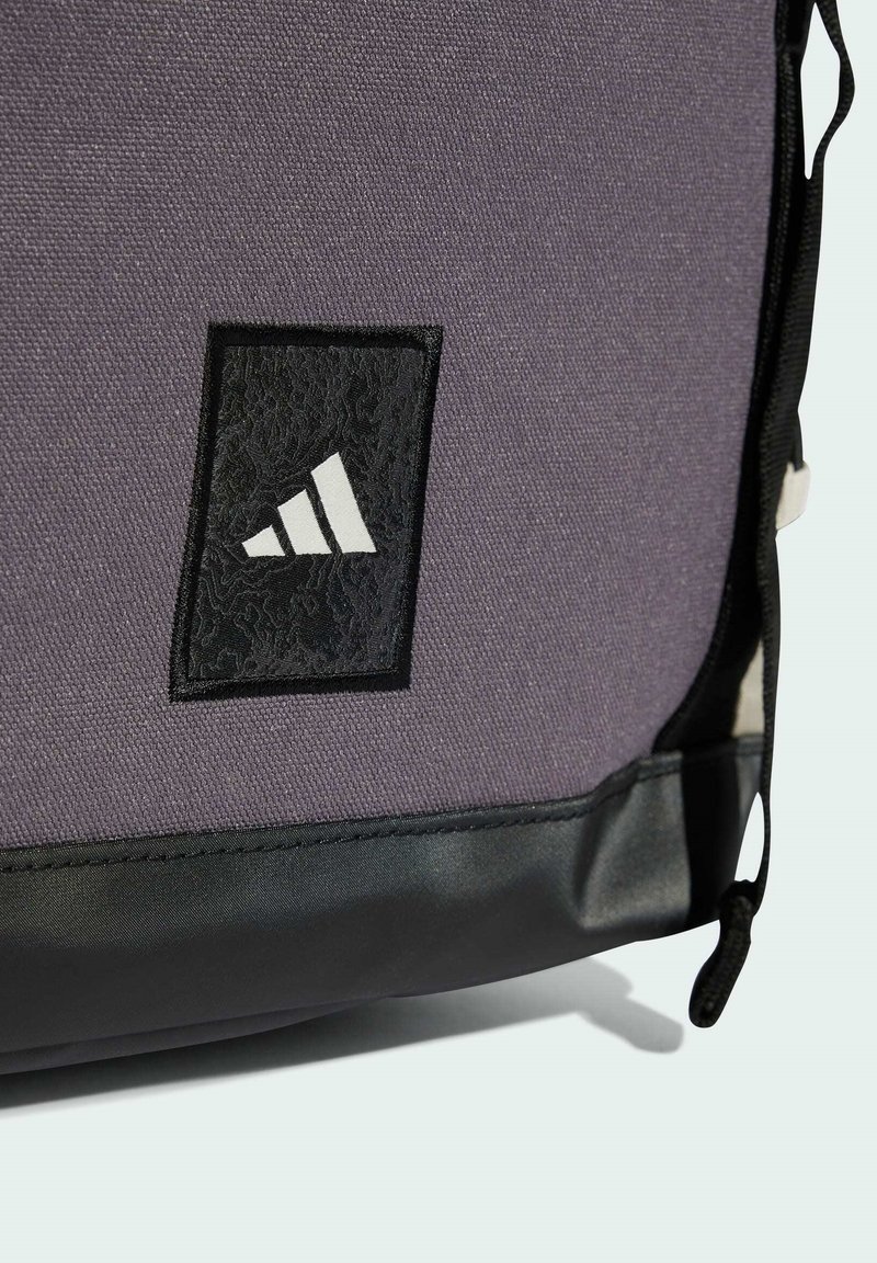 Lila Stofftasche mit schwarzem Bodensaum. Merkmale ein strukturiertes schwarzes Etikett mit weißem Logo und vertikaler Kordelzugverschluss.