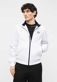 Νέος με σγουρά μαλλιά, φοράει λευκό μπουφάν με φερμουάρ EA7 Emporio Armani πάνω από μαύρο T-shirt με λογότυπο EA7 και μαύρο παντελόνι.