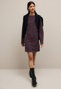 Robe à manches courtes avec un fond bleu-vert, ornée d'un motif d'arbres rouges et verts. Portée avec un châle d'épaule en maille bleu foncé. Bottines noires.