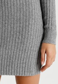 Grå stickad klänning med vertikal ribbning, med lång ärmar och en nederkant som sträcker sig till mitten av låret, med en liten logotypdetalj nära botten.