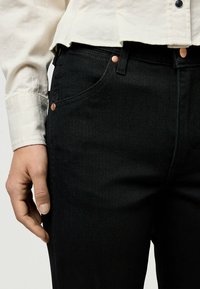 Jeans de mezclilla negra con corte ajustado, que presentan herrajes en tono cobre, bolsillos frontales y un sutil detalle de costura a lo largo de las costuras.