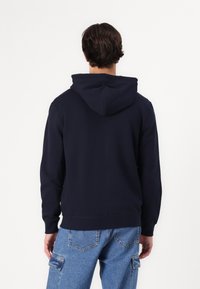 Sweat-shirt à capuche bleu marine en tissu coton mélangé, doté d'une poche kangourou et de poignets côtelés, porté sur un denim bleu clair.