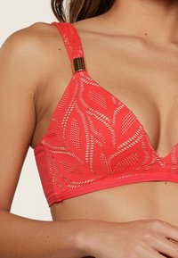 Koraalgekleurde lace bralette met triangulaire cups, voorzien van bladenmotieven, textuurstof en een metalen accent op de band. Gladde onderband.