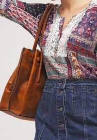 Borsa tote in pelle marrone con finitura testurizzata. L'outfit presenta una blusa a fantasia multicolore e una gonna di jeans con dettagli sui bottoni.