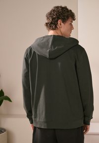 Donkergroene hoodie met een trekkoordcapuchon, zachte textuur, lange mouwen en een relaxte pasvorm, getoond vanuit de rug tegen een neutrale achtergrond.