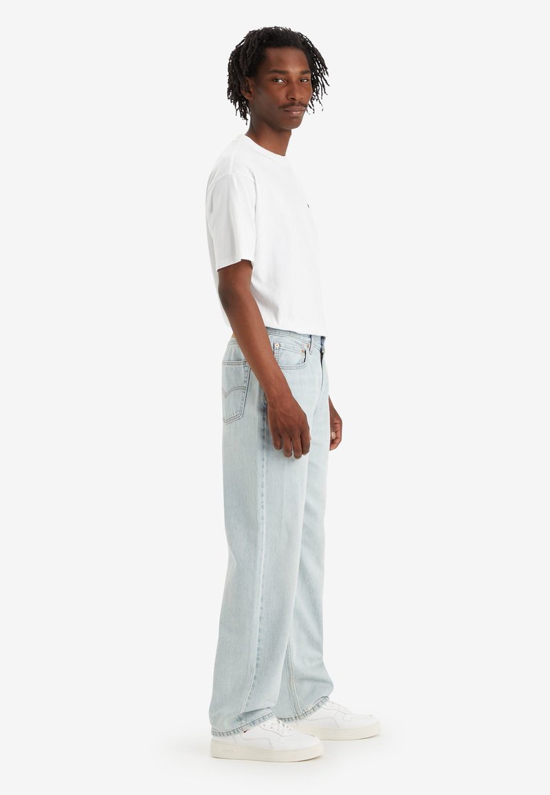 Jeans de mezclilla de corte relajado en azul claro combinados con una camiseta blanca de manga corta. Presentan un logotipo sutil y zapatillas blancas.