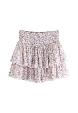 Nuori tyttö hymyilee, pukeutuneena vaaleanpunaiseen perhost-paitaan, kukallinen kerroksellinen hame, vaaleanpunainen lippis ja vaaleanpunaiset tennarit, seisoo yksiväristä taustaa vasten.