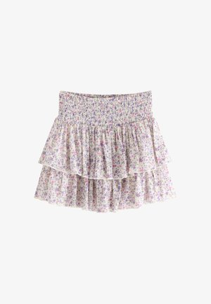 Ung pige smilende, iført pink t-shirt med sommerfugl, blomsteret lagdelt nederdel, pink kasket og pink sneakers, stående mod en ensfarvet baggrund.