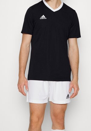 Mann trägt schwarzes V-Ausschnitt Adidas-Sportshirt und weiße Adidas-Shorts, steht vor einem einfarbigen hellen Hintergrund.