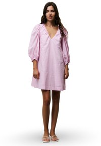 Rotes Baumwollkleid mit tiefem V-Ausschnitt, Puffärmeln und kniehoch geschnittenem Saum. Glatte Textur mit subtiler Schimmer. Weiße Sandalen mit Riemchen.