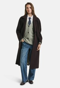 Lungo cappotto in lana marrone scuro, sovrapposto a un cardigan verde, una camicia bianca e jeans a gamba larga blu. Un paio di scarpe in pelle marrone completano l'outfit.