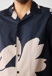 Camisa de manga corta en tejido azul marino con un gran patrón floral beige, que presenta un cuello tipo campamento con muesca y botones negros en el frente.