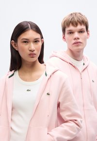 Ροζ φούτερ με φερμουάρ και τσέπες καγκουρό, πράσινες λεπτομέρειες και κέντημα με λογότυπο. Τα μοντέλα φορούν λευκά μπλουζάκια από μέσα. Απλός, καθαρός σχεδιασμός.