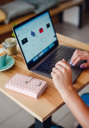 Light pink wallet with dark polka dots and silver hardware on a wooden table beside a laptop. Hand visible typing on keyboard.