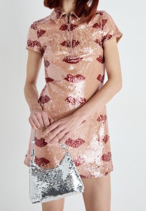 Vestido de lentejuelas en rosa con estampados de labios rojos, con un cuello con cierre de cremallera. Sosteniendo un bolso pequeño de lentejuelas plateadas con un diseño curvo.