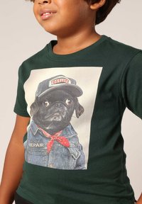 Groene katoenen t-shirt met een grafisch ontwerp van een hond in een denim jacket met een rode bandana, die een marineblauw petje draagt met het label "DEELUXE".