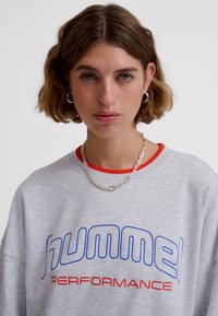Grå sweatshirt med texten "hummel PERFORMANCE" i blått och rött, med rund halsringning med orange detalj och en mjuk textur.