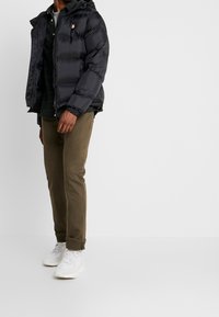 Chaqueta negra acolchada con capucha, con un emblema en el pecho, combinada con pantalones verde oliva y zapatillas blancas. Diseño y textura simples.