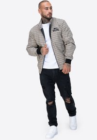 Beżowa i czarna kurtka bomberka w kratę, z ribbowanymi mankietami i zamkiem błyskawicznym z przodu. Noszona z przetartymi czarnymi dżinsami i białymi trampkami.