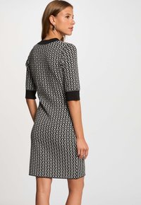Frau in einem knielangen Kleid mit schwarz-weißen geometrischen Mustern und Ellbogenärmeln, die im Profil vor einem schlichten Hintergrund steht.