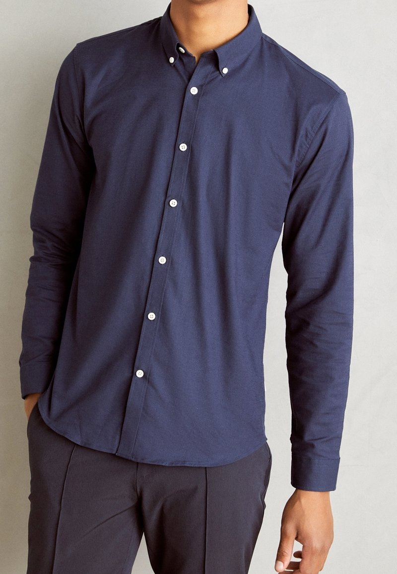 Homme portant une chemise cintrée bleu marine à manches longues boutonnée avec des boutons blancs et un pantalon foncé, debout devant un fond uni.