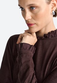Brown long-sleeve top with ruffled cuffs, made of soft fabric. Model has hand on chin, displaying silver drop earrings.