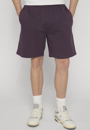 Muž v tmavofialových kraťasoch, bielych ponožkách a bielych teniskách New Balance, stojaci s rukami v kapse na jednofarebnom pozadí.