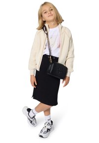 Crème ribbel ritsjack, witte graphic t-shirt, zwarte rok, zwarte schoudertas, witte sneakers met zwarte accenten en gebrandmerkte witte sokken.