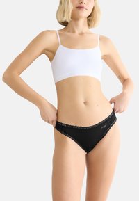 Vrouw in een witte cropped camisole en zwart kantafgebiesde bikini-onderbroek, die de tailleband met beide handen aanpast.