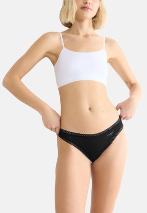 Vrouw in een witte cropped camisole en zwart kantafgebiesde bikini-onderbroek, die de tailleband met beide handen aanpast.