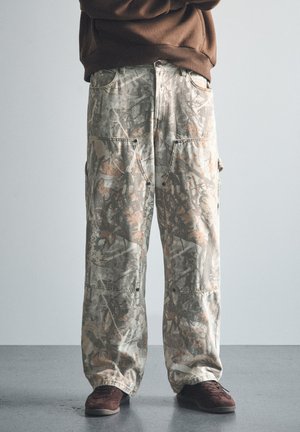 Personne portant un pantalon camouflage à jambes larges et des baskets marron foncé, debout devant un fond uni, les bras croisés.