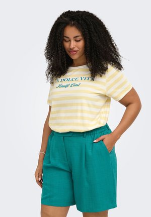 Femme aux cheveux bouclés portant un t-shirt rayé jaune et blanc avec le texte "A Dolce Vita Amalfi Coast" et un short taille haute couleur sarcelle, main dans la poche.