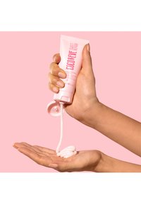 Roze tube van haarbehandeling vastgehouden boven een palm, waarbij een romig, wit product wordt gedoseerd. De achtergrond is effen roze, wat het product benadrukt.