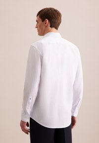 Witte blouse met lange mouwen en subtiele lichtblauwe strepen, met knoopmanchetten en een afgeronde zoom. Achteraan is een gladde, lichte stof zichtbaar.