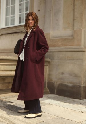 Femme debout à l'extérieur portant un long manteau bordeaux, une chemise blanche, un pantalon foncé et des chaussures claires près d'un bâtiment en pierre avec des fenêtres.